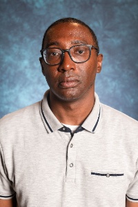 Picture of Cecil Rose, Director of Black Programs  The image is a portrait of a person taken against a blue mottled background. He has a neutral expression and is wearing rectangular glasses with dark frames. His hair is short and he is dressed in a light gray polo shirt with a collar and short sleeves. The polo shirt features dark blue trim along the collar and sleeve edges, two buttons on the placket, and a small pocket on the left side of the chest with the same dark blue trim and a button. The background provides a soft contrast to the subject, allowing him to stand out clearly in the image.